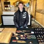 Jaxon Bourne sits in his booth at the Homer Farmers Market with his handmade knives, pens and cutting boards on Saturday, Sept. 13, 2025, in Homer, Alaska. Photo by Christina Whiting